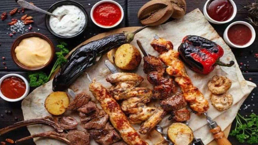 Vista cenital de una tabla de madera con varias brochetas de carne a la parrilla, verduras, pan plano y varias salsas en cuencos pequeños.