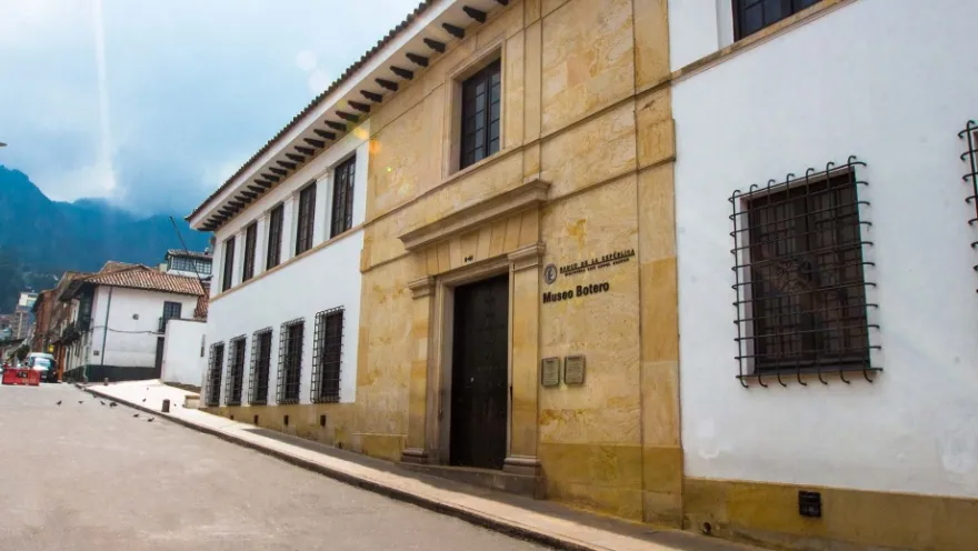 Side view of the Botero Museum's facade. It's a two-story building made of stone and concrete, painted white. A sign above the entrance reads: "Botero Museum."