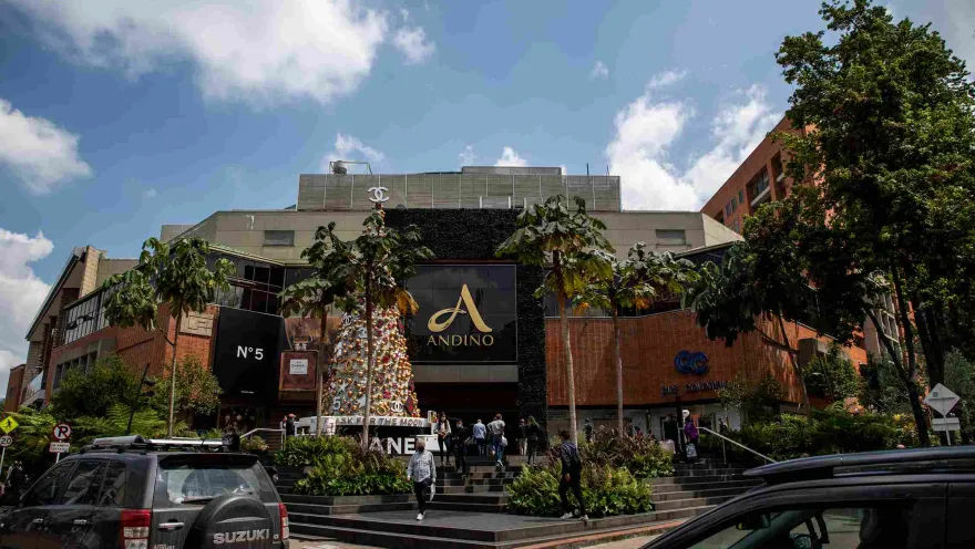 Facade of a building with at least three stories, made of brick and concrete, featuring a large, gold letter A and the word ""Andino."" In front, there are stairs with people and a few cars nearby.