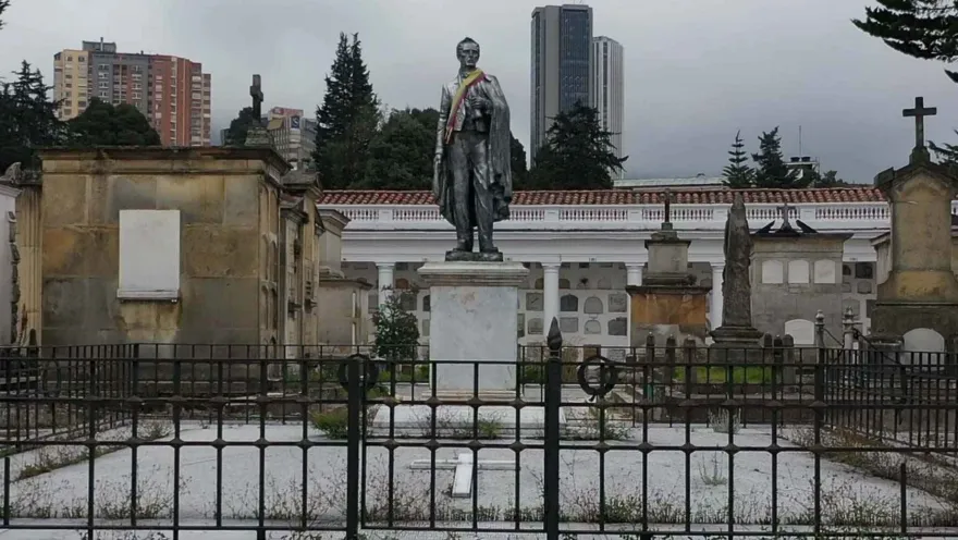 Stone statue in a mausoleum, seemingly of General Santander, with a ribbon in the colors of the Colombian flag. Several other stone mausoleums are nearby. Buildings can be seen in the distance, above the mausoleum.