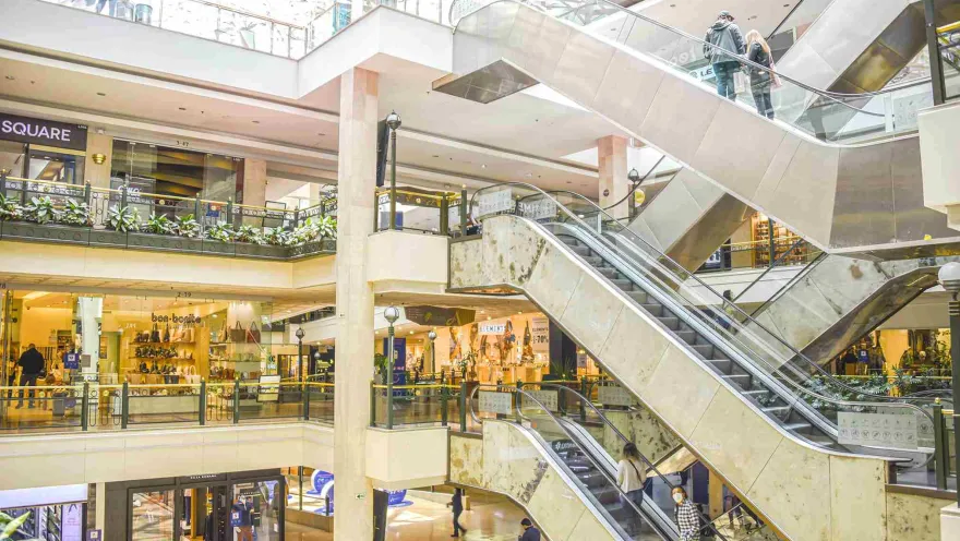Interior of a shopping center showing escalators, shops on several floors, and a few people.