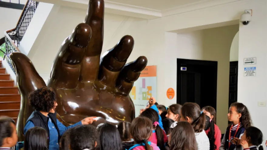 A giant, brown, hand-shaped sculpture surrounded by children and an adult woman who appears to be a guide or teacher.