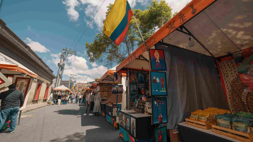 A street lined with vendor stalls displaying handicrafts and paintings, with people browsing in some of them. A Colombian flag hangs prominently on one of the stalls.