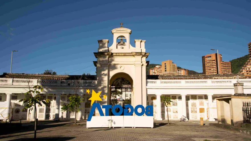 Main entrance of the Central Cemetery, featuring a tall arched gate with a bell tower and clock. At the front is a large structure with the word 'Bogotá' in blue letters and a yellow star above the letter A.