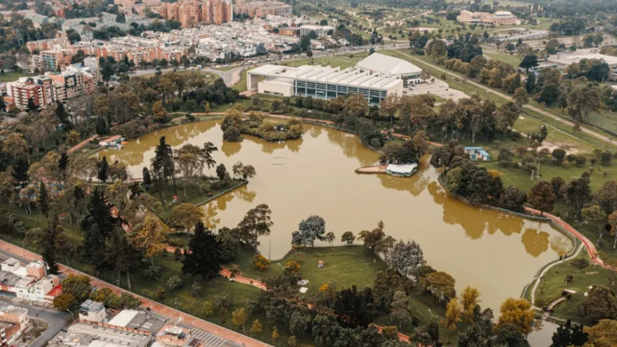 Aerial view of an urban park with a large lake surrounded by trees, pedestrian paths, and green spaces, with city buildings and neighborhoods in the background.