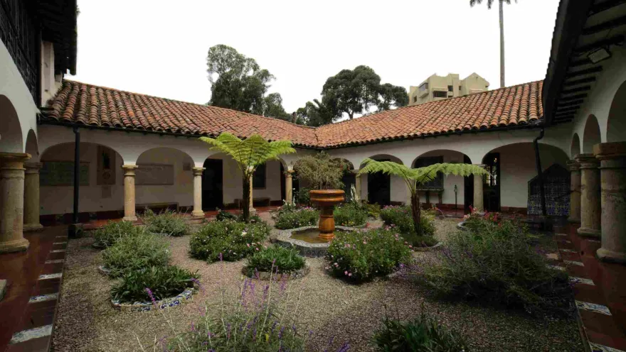 Interior courtyard of a colonial-style building with arched and columned corridors, red tile roofs, and a central garden with plants, ferns, and a small stone fountain.