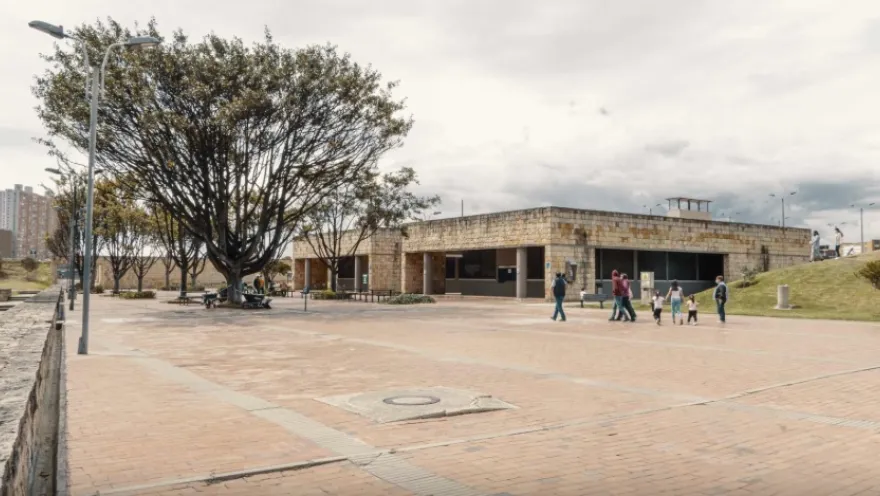 A spacious open-air brick plaza with a low stone building and dark windows in the background. To the left, a large tree provides shade next to benches and lampposts, while several people stroll through the open space under a cloudy urban sky.