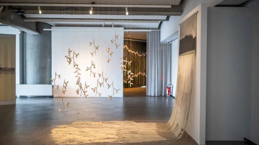 A contemporary art installation in the Bancolombia Art Gallery, where delicate suspended figures hang from the ceiling in front of a white wall, while threads or textile fibers cascade from a structure, forming a kind of canopy on the floor.