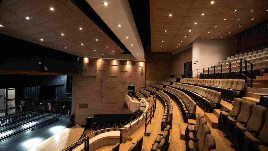 Interior of a modern theater with semicircular tiered seating facing an illuminated stage. The space features warm-toned walls, black railings, and a ceiling with multiple recessed lights.