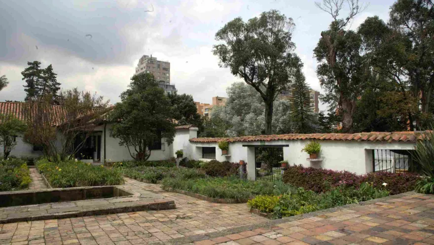 Garden of a colonial house with brick pathways, a small rectangular pond, and white walls with red tiles, surrounded by trees and vegetation, with modern buildings visible in the background.