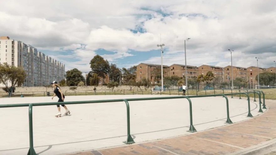 An outdoor skating rink surrounded by green railings, where a person skates on the concrete. In the background, residential buildings, trees, and lampposts are visible under a partly cloudy sky, in a wide-open urban setting.