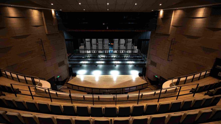 View from the tiered seating of a modern theater with semicircular seating. In the foreground, the illuminated stage and the technical structure behind the curtain are visible, with warm-toned walls and a ceiling with recessed lights illuminating the auditorium.