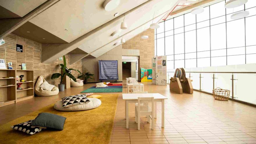 Bright interior space set up for children's activities, with small tables and chairs, cushions on rugs, and shelves with books and materials. The space has large windows that let in natural light and brick walls that create a warm and educational atmosphere.