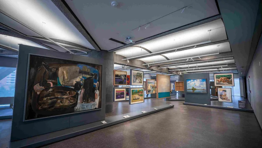 Interior view of the Bancolombia Art Gallery, a modern exhibition space showcasing various large-format paintings and framed works on panels and walls. The spacious room, with lighting directed from the ceiling, allows visitors to explore the exhibition and appreciate the works from different angles in a contemporary setting.