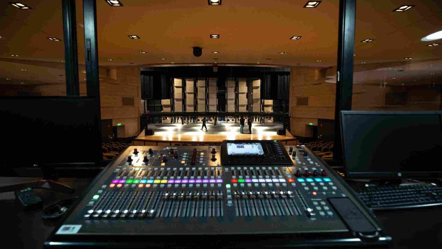 View from the technical booth of a theater, showing a sound console with buttons and colored lights in the foreground. In the background, the illuminated stage and part of the auditorium with rows of seats are visible, while some people walk on the stage during a rehearsal or technical setup.