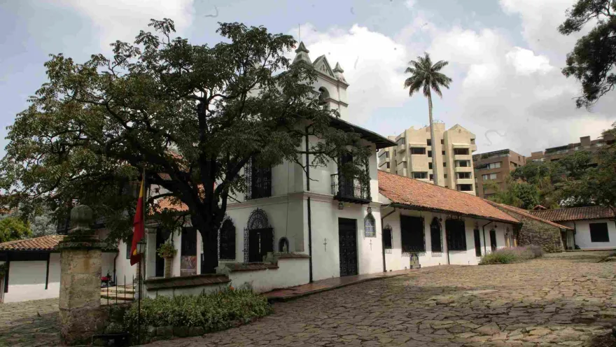 White colonial house with red tile roofs and a small bell tower, surrounded by trees and a cobblestone courtyard, with modern buildings visible in the background.