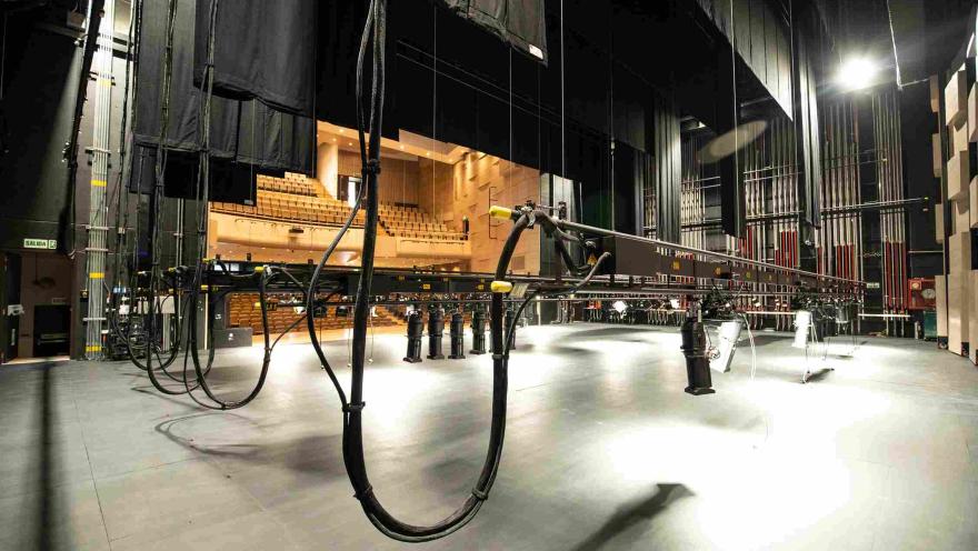 View from the stage of a theater towards the auditorium, showing the tiered seating and the stage's technical infrastructure. In the foreground, cables, lighting rigs, and stage equipment are visible, hanging from the ceiling.