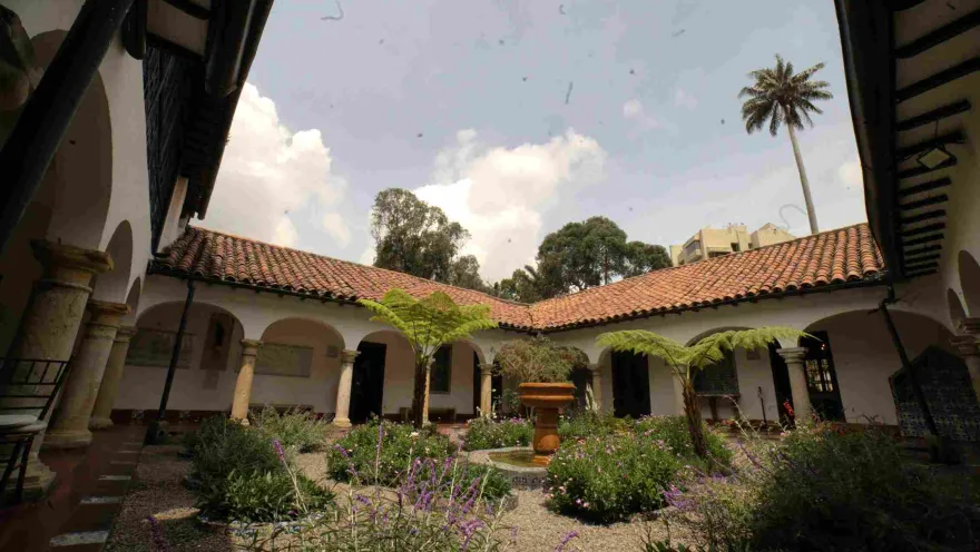 Interior courtyard of a colonial house with arched and columned corridors, red tile roofs and a central garden with plants, flowers and a stone fountain surrounded by vegetation.