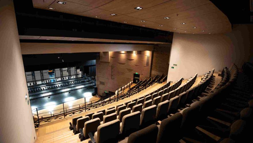 Side view of the interior of a modern theater with rows of cushioned seats in tiered seating that slope down towards the stage. The space features warm ceiling lighting and black railings, while in the background, the stage and its technical structure are visible.