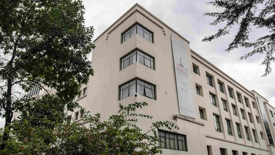 La imagen muestra un edificio de estilo sobrio y elegante, con fachada en tonos claros y grandes ventanas en esquina que aportan modernidad. Está rodeado de árboles y vegetación, lo que le da un ambiente tranquilo.