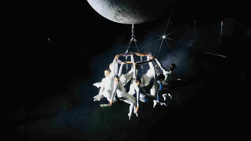 Escena artística en un espacio oscuro: varios acróbatas vestidos de blanco están suspendidos en una estructura colgante bajo una gran luna, iluminados por un foco que resalta la atmósfera teatral y aérea.