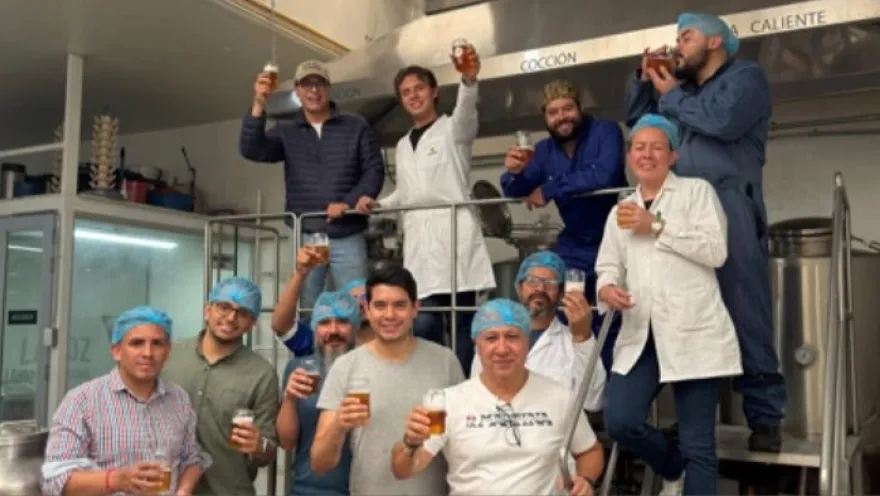 Grupo de once personas en una planta de producción, posando y brindando con vasos de cerveza. La mayoría lleva cofias o gorros de higiene y algunos visten batas blancas, lo que sugiere un entorno de elaboración de alimentos o bebidas.
