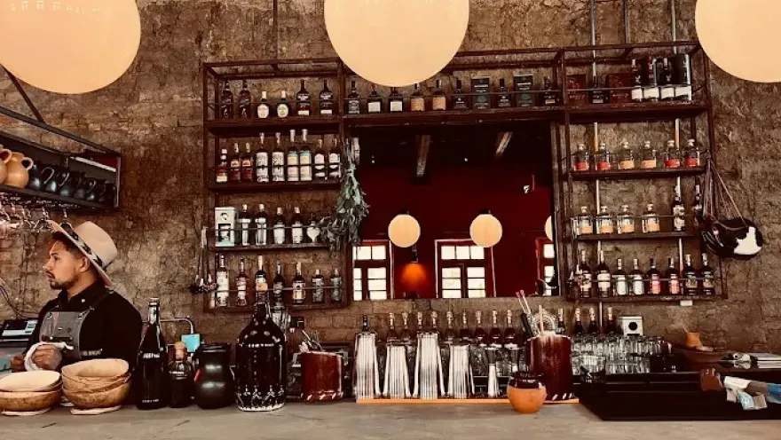 Barra de un bar con estilo rústico e industrial, con paredes de ladrillo y estanterías metálicas llenas de botellas de licor y frascos. Sobre la barra de madera se ven utensilios de coctelería, vasos y recipientes. Tres lámparas redondas cuelgan del techo, iluminando el espacio con luz cálida.