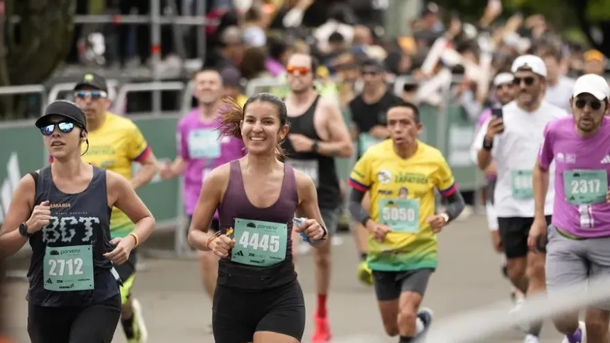 Grupo de corredores participa en una carrera, con una mujer sonriente en primer plano y público animando al fondo.