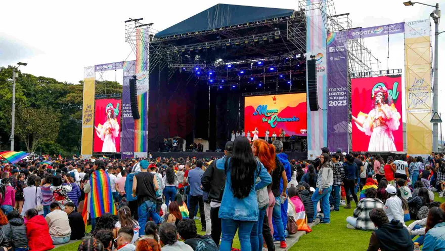 Multitud de personas reunidas frente a un gran escenario al aire libre durante un evento del orgullo LGBTI+ en Bogotá; en las pantallas se ve una artista cantando, mientras asistentes llevan banderas arcoíris y observan el espectáculo sobre el césped.