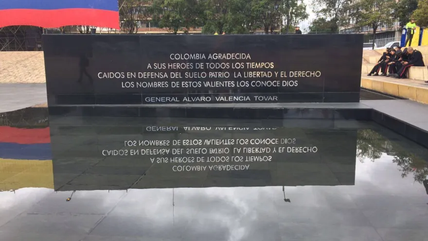  Monumento con placa conmemorativa a héroes caídos, reflejada en una superficie de agua, con bandera de Colombia al fondo.