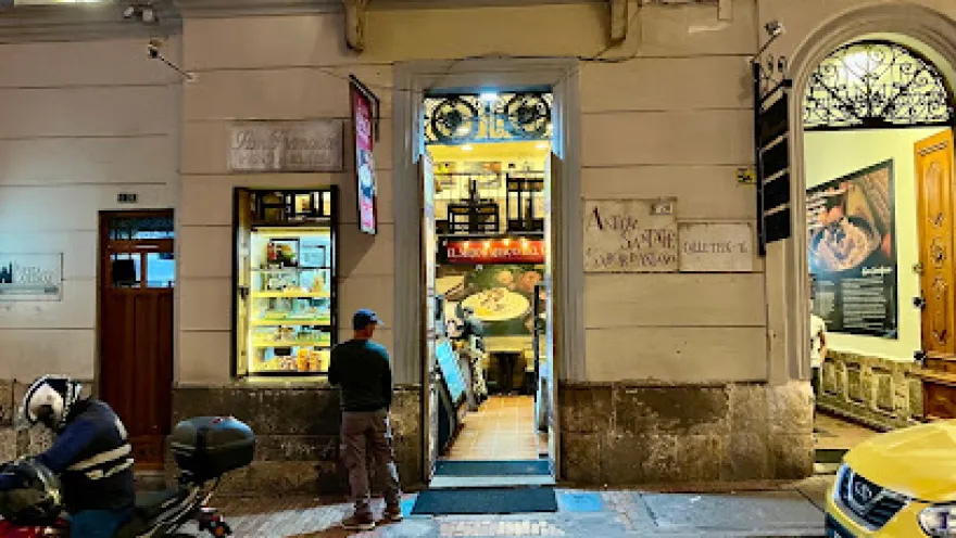 Fachada nocturna de un restaurante tradicional en una calle del centro histórico. La puerta abierta deja ver el interior iluminado con mesas dispuestas para los comensales, mientras una vitrina exhibe productos o alimentos. Afuera, una persona observa el lugar y a un costado hay motocicletas estacionadas y un taxi amarillo, típico de la ciudad.