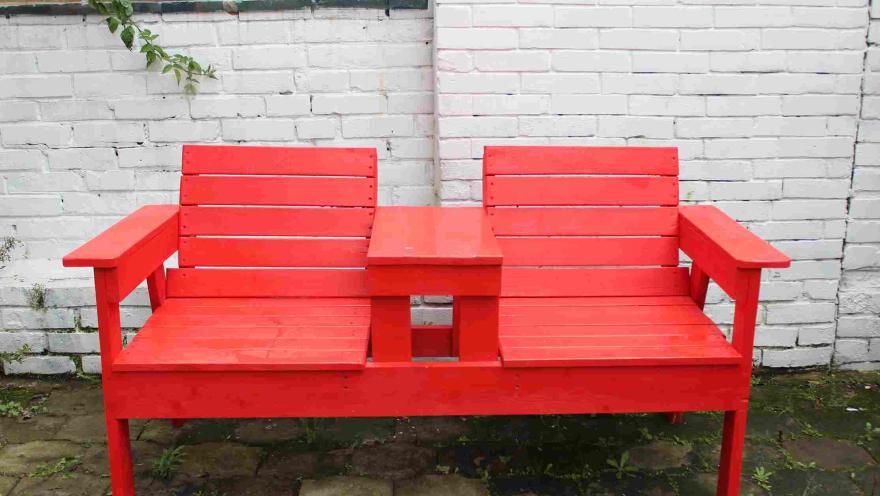 Red double bench with a central table, in front of a white brick wall.