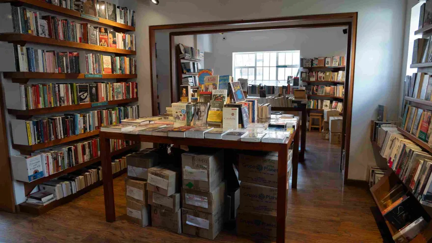  Interior de una librería con estanterías llenas de libros, una mesa central de exhibición y cajas de ejemplares apiladas debajo.