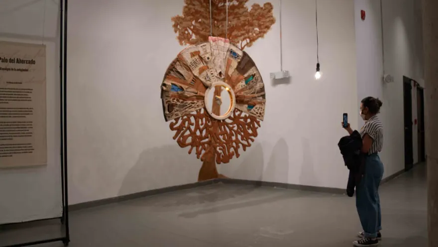 Persona observa y fotografía una obra artística colgante con forma de árbol en una sala de museo.