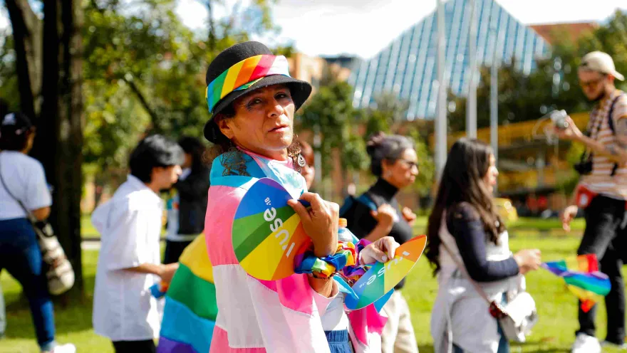Persona en un parque durante una marcha del orgullo LGBTI+, con sombrero negro adornado con una cinta arcoíris y una bandera multicolor sobre los hombros; sostiene abanicos con colores de la diversidad mientras otras personas caminan alrededor en un ambiente soleado.