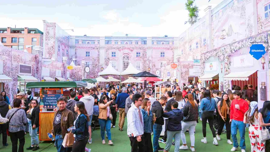  Multitud de personas recorre una feria al aire libre con puestos de comida y marcas, en un espacio decorado con fachadas rosadas y ambiente festivo.