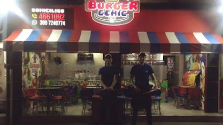  Fachada de un restaurante llamado “Burger” iluminada de noche, con un toldo a rayas rojas, blancas y azules. En la entrada, dos empleados con uniforme oscuro están de pie frente a parrillas, atendiendo al público.