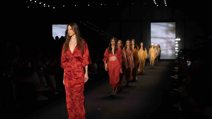 Fila de modelos desfila en pasarela con vestidos en tonos rojos y estampados, ante público en un evento de moda.