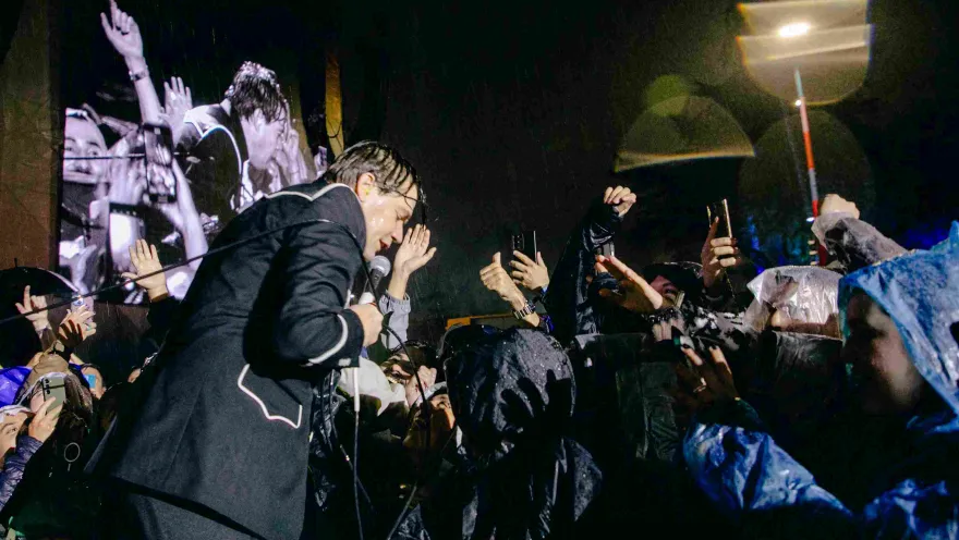 A singer approaches the audience in the rain, who raise their hands and cell phones while enjoying the concert.