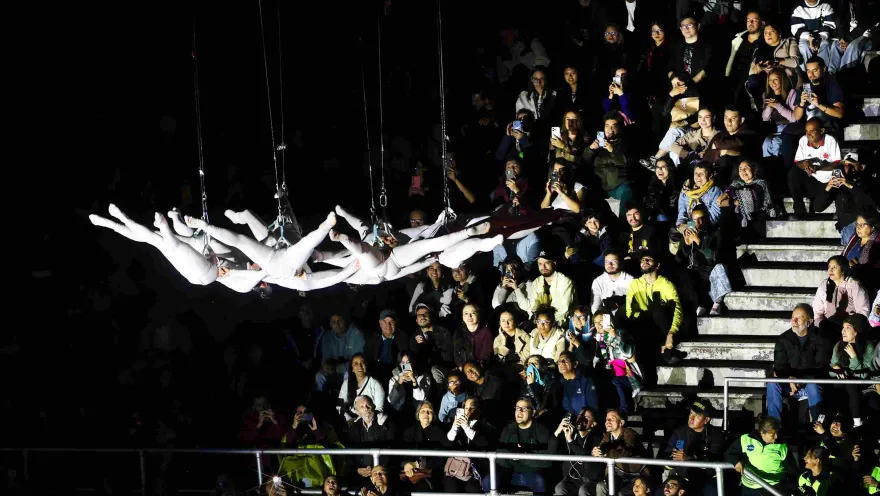  Espectáculo en vivo con acróbatas vestidos de blanco suspendidos horizontalmente en el aire, mientras un público numeroso observa desde gradas, muchos grabando con sus celulares en un ambiente nocturno.