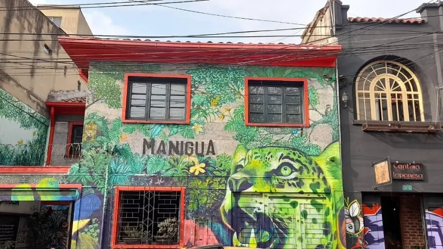  Pasillo interior de un bar o café alternativo con iluminación en tonos azules y morados. Las paredes están cubiertas de carteles, ilustraciones y grafitis, creando un ambiente urbano y artístico. A ambos lados, grupos de personas conversan sentadas en sillas y mesas pequeñas, algunas con bebidas.