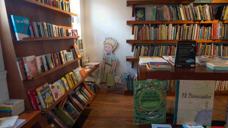 Sección infantil de una librería con estanterías de madera llenas de libros ilustrados, una mesa de exhibición y una figura de El Principito en el fondo.