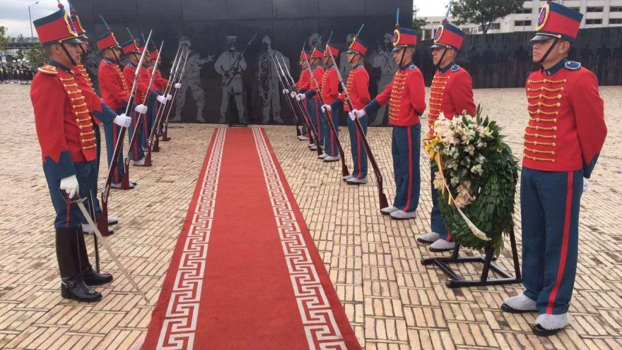  Guardia de honor con uniformes rojos custodia un monumento, junto a una ofrenda floral y una alfombra roja.
