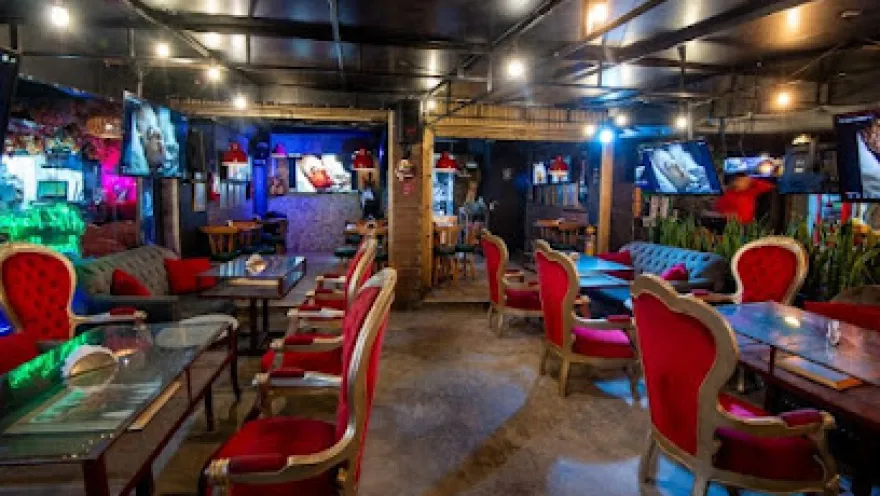 Interior de un restaurante de estilo moderno con iluminación cálida y toques de neón. El espacio cuenta con mesas de madera y sillas tapizadas en rojo con diseño clásico, organizadas en filas. En las paredes y columnas hay pantallas de televisión encendidas y elementos decorativos contemporáneos.