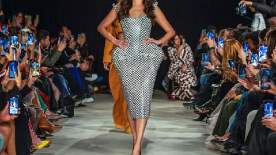  Modelo desfila en pasarela con vestido ajustado a cuadros, mientras el público toma fotos a ambos lados.