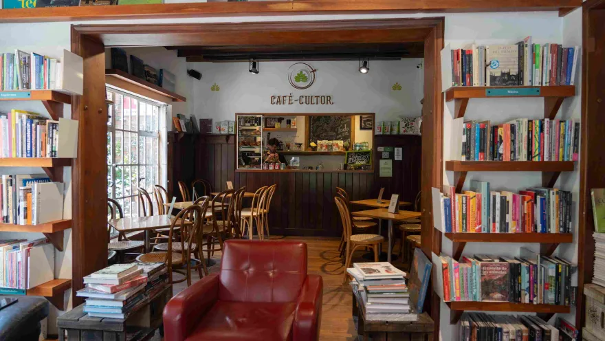 Interior de una librería con café, rodeada de estanterías de libros, mesas de madera y un mostrador al fondo bajo el letrero “Café-Cultor”.