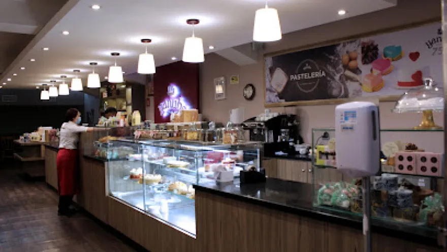 Interior de una pastelería moderna y bien iluminada, con vitrinas de vidrio que exhiben una variedad de postres y productos de repostería. El espacio cuenta con un largo mostrador de madera, lámparas colgantes de luz blanca y paredes decoradas con imágenes de dulces.
