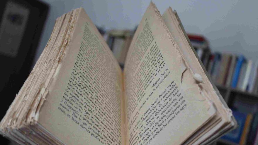 An open antique book with worn pages and visible text, set against a casual background of bookshelves.