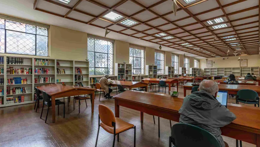 La imagen muestra una sala de lectura amplia y bien iluminada, con mesas de madera y estanterías llenas de libros a lo largo de las paredes. Varias personas leen o trabajan en silencio, creando un ambiente tranquilo y concentrado. La luz natural que entra por las ventanas y el techo geométrico aportan calidez y orden al espacio.