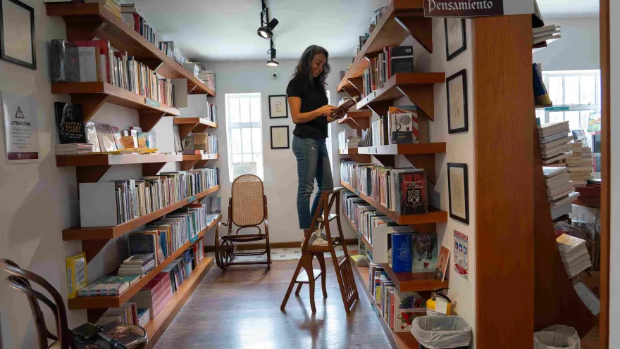 Mujer sobre una escalera leyendo un libro entre estanterías de madera llenas de libros en una librería.